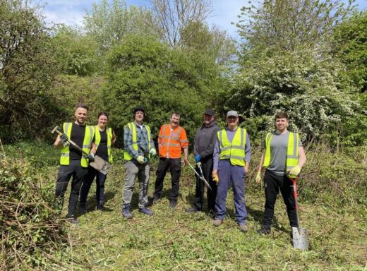 A group of six people in high-visibility vests holding gardening tools in a green area.