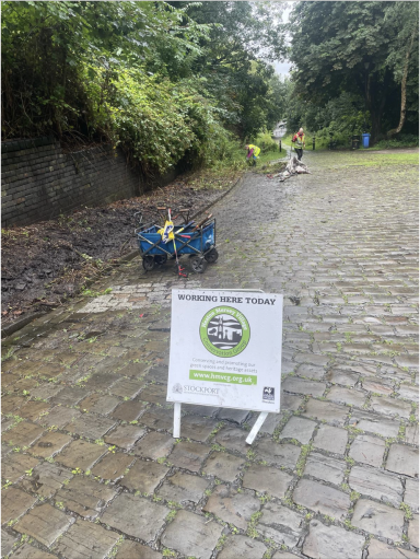 Sign on a path indicating a community clean-up event, with volunteers visible in the background.